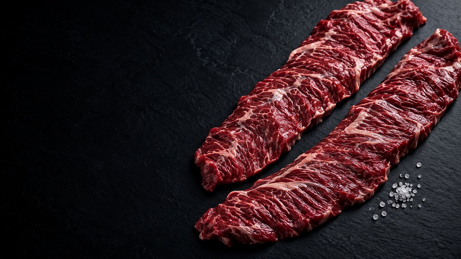 Two raw, well-marbled inner skirt steaks on dark slate with coarse sea salt